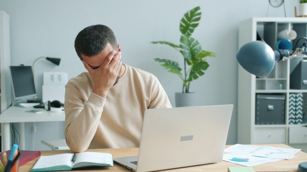 Müder Mann sitzt mit Laptop am Schreibtisch und hält sich das Gesicht