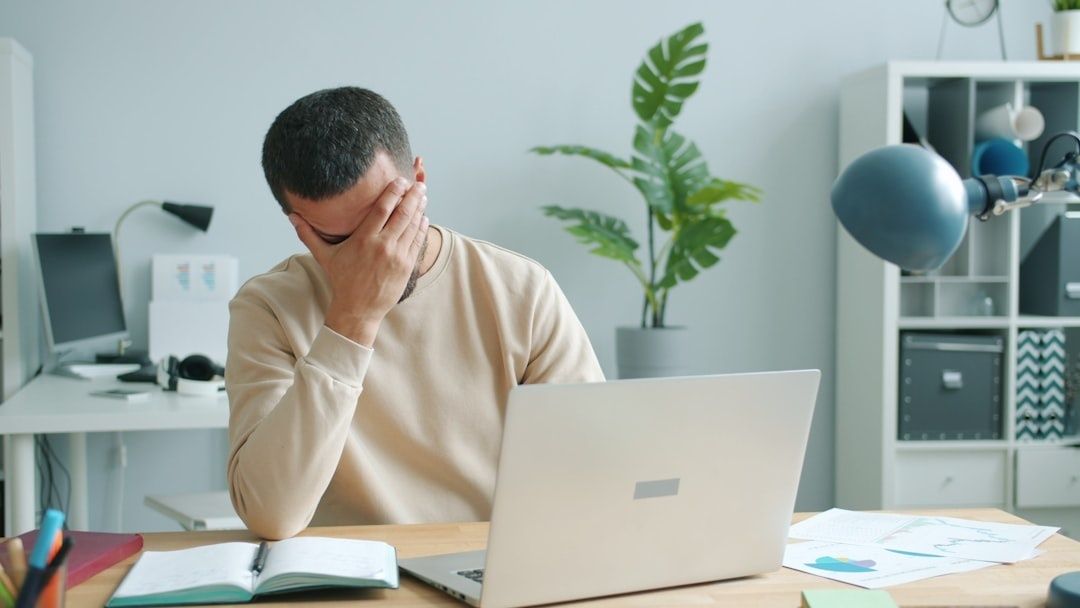 Müder Mann sitzt mit Laptop am Schreibtisch und hält sich das Gesicht